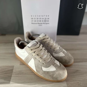 Maison Margiela sneakers i mocka och skinn - Snygga sneakers från Maison Margiela i beige mocka och vitt skinn med klassisk gummisula. Låg modell med rund tå och snörning. Perfekta för dig som gillar stilrena och trendiga skor med lyxig känsla.
