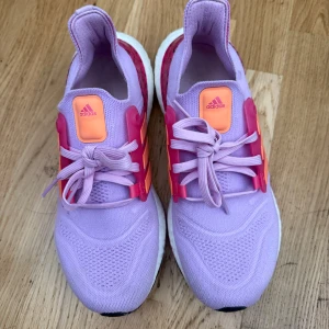 Lila Adidas Ultraboost sneakers - Säljer ett par Adidas Ultraboost sneakers i lila med rosa och orange detaljer. Skorna har stickad ovandel, snörning och vit Boost-sula för extra komfort. Snygga kontraster med rosa och orange på sidorna och hälen. Perfekta för dig som vill sticka ut med färg på fötterna!