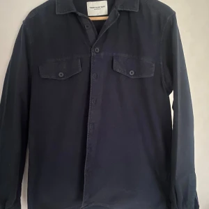Mörkblå overshirt från These Glory Days - Stilren mörkblå overshirt från These Glory Days med klassisk krage, två bröstfickor med knappar och långa ärmar. Perfekt att bära öppen över en t-shirt eller stängd som skjorta. Gjord i mjuk bomull för en skön känsla.