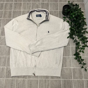 Ljusgrå kofta från Polo Ralph Lauren - Snygg ljusgrå kofta från Polo Ralph Lauren med klassisk broderad logga på bröstet. Tröjan har hel dragkedja, ribbade muddar och krage med svarta och vita ränder. Tillverkad i mjuk bomull, perfekt för lager-på-lager och chill stil.