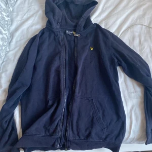 Mörkblå hoodie från Lyle & Scott - Mörkblå hoodie från Lyle & Scott med dragkedja och huva. Klassisk gul logga broderad på bröstet och två fickor framtill. Perfekt för dig som gillar en enkel och stilren look. Tillverkad i mjuk bomull för extra komfort.