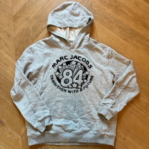 Marc Jacobs hoodie - Säljer en grå hoodie från Marc Jacobs med svart tryck framtill med texten '84 Tradition With A Future' och stjärndetaljer. Använd fåtal gånger. Riktigt skönt och mjukt bomulls material. Den är slutsåld. Storlek 162, men passar även XS. Tröjan är tunn, perfekt för chill dagar. Pris kan diskuteras