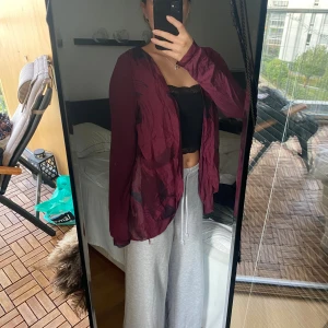 Vinröd omlottkofta i tunn polyester - Säljer en vinröd omlottkofta i tunn, lätt och lite glansig polyester. Koftan har lång ärm, öppen framsida och cool omlottdetalj i ryggen. Perfekt att slänga över ett linne för en chill och snygg look.