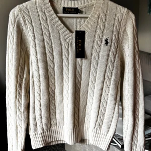 Vit kabelstickad tröja Polo Ralph Lauren - Kabelstickad vit tröja från Polo Ralph Lauren med klassisk v-ringning och broderad logga på bröstet. Tröjan har lång ärm, ribbade muddar och är gjord i mjuk bomull. Perfekt för lager-på-lager och snygg till jeans eller kjol.