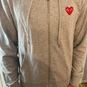 Grå hoodie Comme des Garçons Play - Grå hoodie från Comme des Garçons Play med dragkedja, huva och snörning. Storlek M men passar S. Helt oanvänd