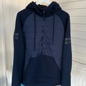 Marinblå hoodie Sail Racing half zip - Snygg marinblå hoodie från Sail Racing med quiltad front, half zip och huva med snörning. Stora loggor på ärmarna och bak på huvan. Perfekt för kyliga dagar, med praktisk känguruficka med dragkedjor. Sportig och modern look.