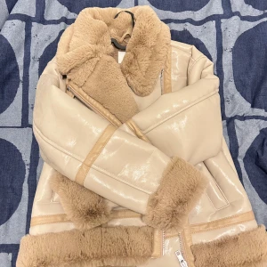 Beige teddyjacka med dragkedja - Säljer en beige teddyjacka från H&M med fluffigt foder och krage. Jackan har glansig utsida, breda muddar och dragkedja framtill. Perfekt för kalla dagar och ger en cool look med sin oversized passform och mjuka material.