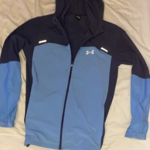 Blå vindjacka från Under Armour - Snygg tvåfärgad vindjacka från Under Armour i marinblått och ljusblått. Jackan har huva, hel dragkedja och reflekterande detaljer. Perfekt för träning utomhus och har logga både fram och bak. Elastiska muddar med diskret logotyp på ärmsluten.