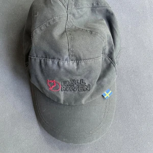 Svart Fjällräven keps med snörning - Svart keps från Fjällräven med broderad logga i rött och svart framtill samt svensk flaggdetalj. Insidan har quiltad svart polyester och röd fleece för extra värme. Justerbar snörning baktill med metallöglor och lås. Perfekt för kyliga dagar utomhus.