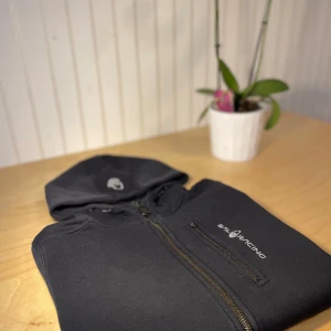 Svart hoodie från Sail Racing - Svart hoodie från Sail Racing med dragkedja framtill och en bröstficka med dragkedja. Tröjan har broderad logga på huvan och bröstet. Mjuk och skön, perfekt för kyliga dagar. Klassisk passform och långa ärmar.