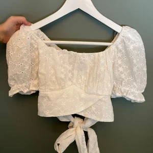 Vit sommar topp - Superfin vit blus med broderade blommor och puffiga korta ärmar. Toppen har omlottmodell med knyt baktill och är croppad. Perfekt för en somrig look med en jeanskjol🩷