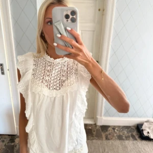 Vit blus med spets och volanger - Säljer en superfin vit blus med spetsdetaljer över bröstet och volanger längs axlarna. Blusen är ärmlös och har en romantisk vibe med broderade mönster och rund hals. Perfekt för dig som gillar en feminin och söt stil.