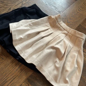 Beige kortkjol och en svart kortkjol med veck från Zara  - Superfin beige kortkjol från Zara med snygga markerade veck som ger extra volym. Kjolen har en klassisk midja och är i ett mjukt, lätt material som känns skönt mot huden. Perfekt för dig som gillar en stilren och trendig look.