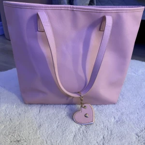 Rosa Väska  -  rosa väska med långa axelband och en  hjärtformad nyckelring i guld och rosa. Väskan är rymlig och har ett prickigt innerfoder i ljusrosa. 