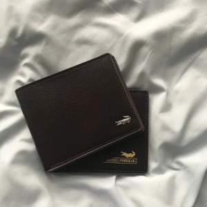 Lacoste wallet - Den är helt ny och oanvänd och snygg mörkbrun plånbok i skinn från Lacoste med stilren design. Den fraktas inom 24h. Nytt pris 1000kr