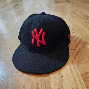 Svart New Era keps med röd NY-logga - Säljer en svart New Era 59FIFTY keps med röd broderad New York Yankees-logga framtill och MLB-logga bak. Klassisk rak skärm och snygg kontrast mellan svart tyg och röda detaljer. Perfekt för dig som gillar streetstyle och sportig look.