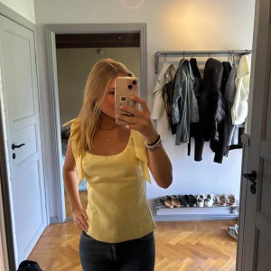 Gul linnetopp med rosettband - Snygg ljusgul linnetopp med breda axelband som knyts till rosetter. Toppen har en rak, figurnära passform 