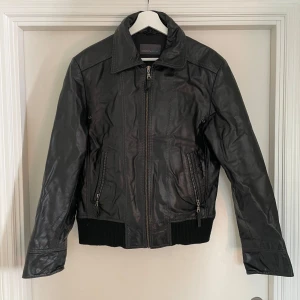 Svart skinnjacka från Rock'n Blue - Snygg svart skinnjacka från Rock'n Blue med klassisk krage, dragkedja framtill och ribbade muddar i midjan. Jackan har två fickor med dragkedja och stilrena sömdetaljer. Perfekt för dig som vill ha en tidlös och cool look.