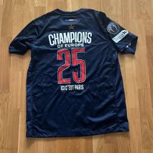 PSG t-shirt - Snygg PSG fotbollströja med mörkblå bas, röd och vit rand längs mitten, och tryckta detaljer som 'Champions of Europe' och nummer 25 på ryggen. 