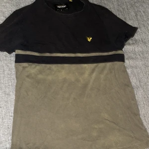 Svart och olivgrön t-shirt Lyle & Scott - T-shirt från Lyle & Scott i svart och olivgrönt med en bred horisontell rand över bröstet. Klassisk rund hals och korta ärmar. Liten gul logotyp på vänster bröst. Mjuk bomullskvalitet, perfekt för chill och streetwear.