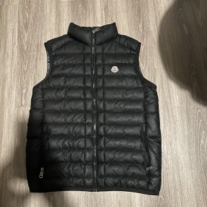 Svart dunväst från Moncler  - Säljer en svart dunväst från Moncler i storlek XXL. Västen har quiltad design, hög krage och dragkedja framtill. Klassisk Moncler-logga på bröstet och två fickor med dragkedja. Perfekt för lager-på-lager och riktigt snygg till streetwear. Den har ett hål i fickan men det är litet 