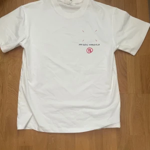 Vit Maison Margiela t-shirt med print - Vit t-shirt från Maison Margiela med svart och röd print fram och bak. Trycket på ryggen har siffror och logga, medan framsidan har diskret Maison Margiela-tryck. Klassisk passform och rund hals. Tillverkad i mjuk bomull. Vid fler frågor eller funderingar är det bara att skriva