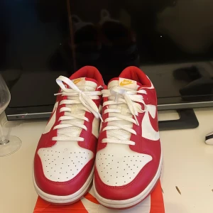 Nike Dunk Low röd/vit - Nike Dunk Low sneakers i rött och vitt med klassisk Swoosh-logga på sidorna. Skorna har vit bas med röda overlays och detaljer, samt gul Nike-brodyr på hälen. Lågt skaft, perforerad tå och vit sula med röd yttersula. Sköna och snygga för dig som gillar streetstyle.