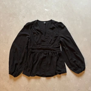 Svart blus med v-ringning från SHEIN - Svart blus från SHEIN med v-ringning och långa puffärmar. Modellen har markerad midja med bred smock baktill och lätt rynkad nederdel. Tunt, flowy tyg som ger en snygg siluett. Perfekt för dig som gillar stilrena och trendiga plagg.