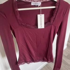 Vinröd långärmad topp från Venderby’s - Supersnygg vinröd långärmad topp från Venderby’s med spetsdetaljer vid halsringning och nederkant. Figurnära passform och fyrkantig urringning ger en trendig vibe. Perfekt att styla med jeans eller kjol för en cool look.