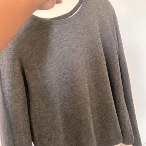 Grå stickad tröja med öppen rygg - Mysig grå stickad tröja med lång ärm och en snygg öppen ryggdetalj. Tröjan har en rund halsringning och ribbade muddar vid ärmslut. 