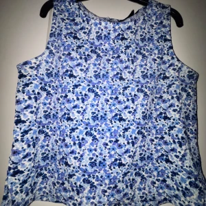 Blommig blå ärmlös linnetopp - Superfin ärmlös topp med blått och vitt blommönster.I storlek 146-152. Toppen har en rak passform och är gjord i ett lätt och luftigt tyg, perfekt för varma dagar. Snygg att styla med jeans eller kjol för en fräsch look.