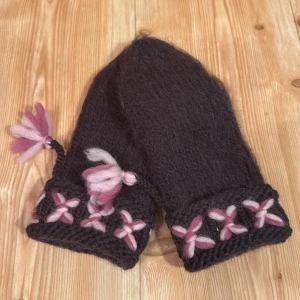 Bruna stickade vantar i lovikkastil - Mysiga bruna stickade vantar med broderade blommor i rosa och ljusrosa runt mudden. Vantarna har en söt tofs i rosa nyanser på sidan. Perfekta för kalla dagar när du vill ha något unikt och färgglatt på händerna. Damstorlek. Handstickade i 100% ull tovade