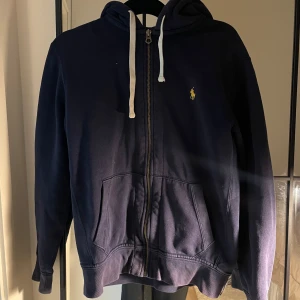 Mörkblå hoodie från Polo Ralph Lauren - Säljer en mörkblå hoodie från Polo Ralph Lauren med dragkedja och huva. Klassisk gul logga på bröstet, vita snören och två stora fickor framtill. Perfekt för dig som gillar en avslappnad och stilren look.