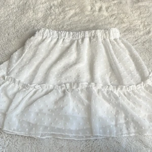 Vit kortkjol med volang från SHEIN - Supersöt vit kortkjol från SHEIN med volangkant och små prickiga detaljer i tyget. Kjolen har resår i midjan för en bekväm passform och är gjord i ett lätt och luftigt syntetmaterial. Perfekt för en fräsch och somrig look!