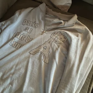 Ljusgrå half zip hoodie med strass - Säljer en ljusgrå hoodie med half zip och strassdetaljer. Framsidan har texten 'paris au clair de lune' och en stor Eiffel-torn design i strass. Mjuk insida och klassisk huva, perfekt för dig som gillar Paris-vibes och bling.