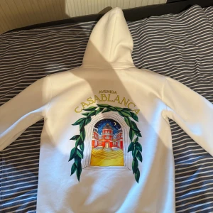 Vit hoodie från Casablanca med tryck - Snygg vit hoodie från Casablanca med färgglatt tryck av en byggnad och gröna blad både fram och bak. Tröjan har huva med snörning och en stor magficka. Perfekt för dig som vill sticka ut med en unik och trendig look.