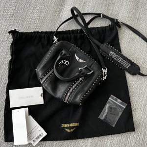 Svart handväska Zadig & Voltaire skinn - Snygg väska från Zadig & Voltaire i skinn med silvriga dragkedjor, nitar och detaljer. Väskan fungerar till allt. Perfekt storlek för det viktigaste och ger en edgy vibe. Den är heller inte så stor så lätt att bära på.
