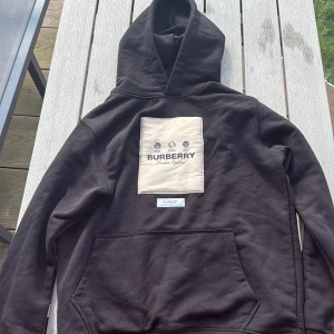 Svart Burberry hoodie med beige patch - Säljer en svart hoodie från Burberry i storlek S. Tröjan har en stor beige patch med Burberry-logga på bröstet och en mindre logga nedanför. Klassisk huva och känguruficka framtill. Perfekt för dig som gillar streetstyle och exklusiva märken.