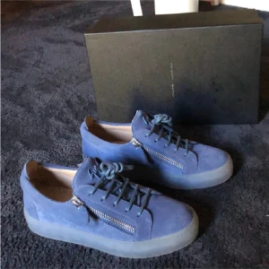 Blå mockasneakers från Giuseppe Zanotti - Snygga sneakers i ljusblå mocka från Giuseppe Zanotti med genomskinlig platt sula, snörning och coola silverfärgade dragkedjor på sidorna. Ikonisk logga på plösen och stilren design som sticker ut.