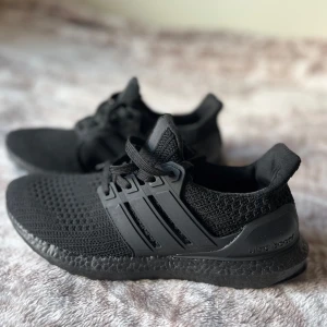 Svarta Adidas Ultraboost sneakers - Säljer ett par helsvarta Adidas Ultraboost sneakers med stickad ovandel och klassiska tre ränder på sidan. Skorna har snörning, platt sula och en riktigt skön boost-dämpning. Perfekta för dig som gillar stilrena och bekväma sneakers till vardags eller träning. HELT NYA-ALDRIG ANVÄNDA