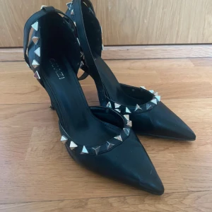 Svarta pumps med nitar och spetsig tå - Svarta pumps med spetsig tå och hög klack. Skorna är dekorerade med silvriga pyramidformade nitar runt foten och hälen för en edgy look. Tillverkade i skinnliknande material och har en elegant siluett som sticker ut.