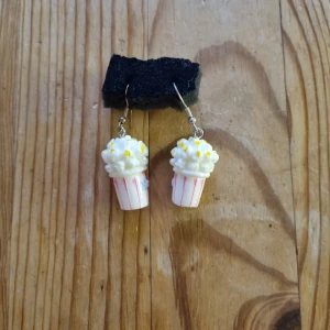 Popcorn örhängen med rosa ränder - Superroliga örhängen formade som popcornbägare med vita och gula detaljer. Bägaren har rosa ränder och popcornet ser fluffigt ut. Perfekt för dig som vill sticka ut och visa din kärlek för snacks! Säljs som ett par.