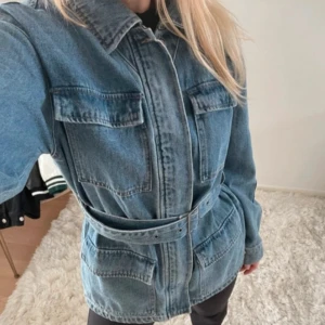 Blå jeansjacka med bälte - Snygg blå jeansjacka med klassisk krage och fyra stora fickor framtill. Jackan har ett matchande bälte i midjan som ger en cool siluett. Långärmad och med knappar längs hela framsidan. Perfekt för dig som gillar avslappnad streetstyle. #hösttrend #denimjacket