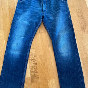 Blå Diesel Krooley jeans regular slim - Säljer ett par blå Diesel Krooley jeans. Klassisk femficksmodell med snygga slitningar och raka ben. Jeansen har tydliga sömmar och coola detaljer på bakfickorna. Perfekta för dig som gillar en avslappnad men stilren look.