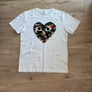 Vit Comme des Garçons t-shirt camohjärta - Vit t-shirt från Comme des Garçons Play med stort hjärta i camouflageprint och broderat rött hjärta med ögon på bröstet. Klassisk passform och rund hals. Perfekt statement-plagg för dig som gillar streetstyle. Den övre ettiketen avklippt.