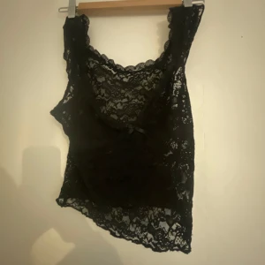Super snygg Svart spetsig linnetopp med rosett - Säljer en helt oanvänd svart ärmlös linnetopp i genombruten spets med söt liten rosett framtill. Toppen har vacker blommig spets och en feminin, croppad passform. Perfekt att styla lager på lager eller som statement-piece till jeans eller kjol.