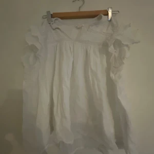 Super fin Vit kortklänning med volang från Gina Tricot - Supersöt vit blus från Gina Tricot med volangdetaljer på ärmarna och luftig passform. Blusen har fina mönster upptill och är perfekt för varma dagar. Materialet känns lätt och fräscht, och modellen är ärmlös med volanger.