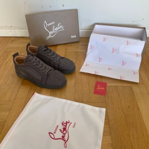 Grå Louboutins - Snygga grå sneakers i mocka från Christian Louboutin med ikoniska röda sulor och coola nitar på tån. Skorna har snörning och en rund tå, perfekt för dig som vill sticka ut med exklusiv streetstyle. Kommer med originalkartong och dustbag.