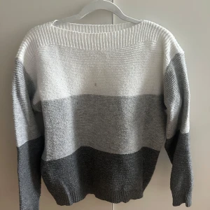 Randig stickad tröja i grått och vitt - Säljer en stickad tröja med breda ränder i vitt, ljusgrått och mörkgrått. Tröjan har en avslappnad passform, rund halsringning och långa ärmar. Perfekt för dig som gillar en enkel men snygg look till höst och vinter. Tyvärr har tröjan en fläck framtil. Tyvärr har tröjan en fläck framtil