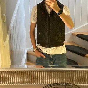 Svart quiltad väst från Polo Ralph Lauren - Quiltad svart väst från Polo Ralph Lauren med orange logga på bröstet. Västen har ribbad krage och nederkant, två fickor med tryckknappar och dragkedja framtill. Perfekt att slänga över en hoodie eller t-shirt för en chill look.